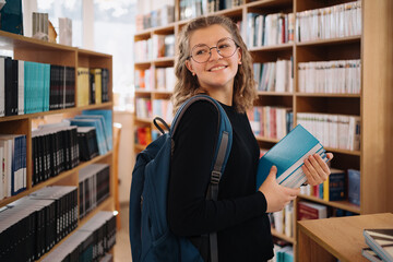 Happy teenage girl or student wearing glasses taking book from shelf in library, wear backpack- People, knowledge, education and school concept