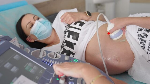 Pregnant woman in mask doing ultrasonography. Doctor checking baby in pregnant woman belly and uses modern technology medical equipment in hospital.