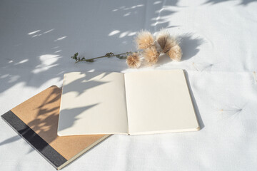 Modern summer stationery still life.Empty diary, notepad mockup with floral element on white linen tablecloth. Long shadows of sunset in the sunlight. Top view