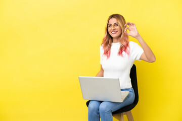 Naklejka premium Young woman sitting on a chair with laptop over isolated yellow background showing ok sign with fingers