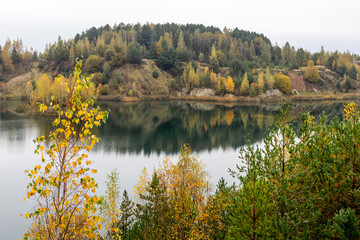 Lake in the autumn landscape. Hilly autumnal mixed forest. with a view through spruce branches and a lone birch tree in the foreground, a yellow and green forest with a mirror image in the water.
