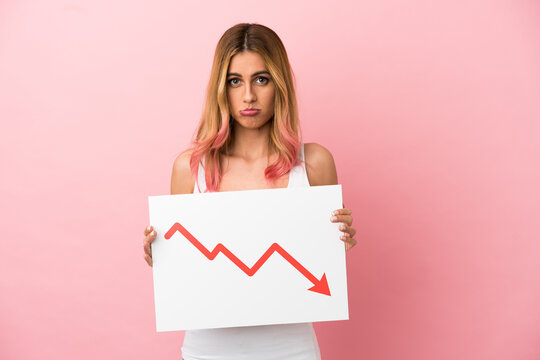 Young Woman Over Isolated Pink Background Holding A Sign With A Decreasing Statistics Arrow Symbol With Sad Expression