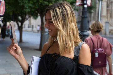 pretty young woman in tourism is looking at a postcard to send to her friends. The girl is carrying a backpack, sunglasses in her hand and a map under her arm. Concept tourism, holidays, travel.