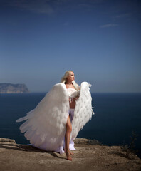 Naklejka premium Young beautiful woman fallen sad angel sits on the sea beach. Creative sexy costume, huge artificial bird wings and white vintage dress. Adult girl with sad face of repentance. Artwork photo