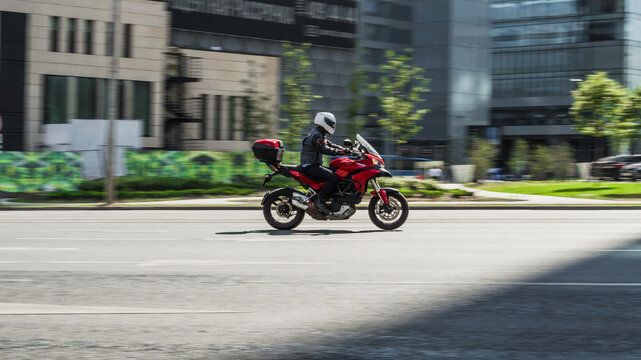 Young man riding fast on red motorbike Ducati. Rider in black clothes and a white helmet, driving on city street on a sunny day