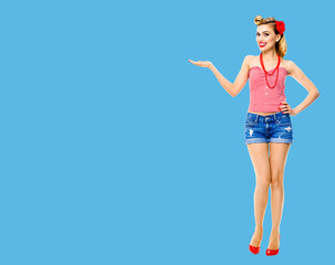 Full body photo - happy excited smiling woman holding, giving, advertising something. Blond girl in pinup style, showing copyspace for text or imaginary. Retro fashion and vintage. Blue background.