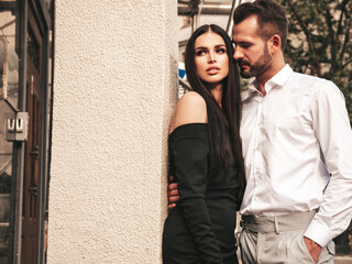 Beautiful fashion woman and her handsome elegant boyfriend in suit. Sexy brunette model in black evening dress. Fashionable couple posing in the street in Europe. Brutal man and his female outdoors