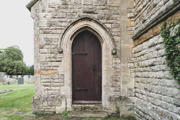 Secondary entrance to a crypt seen at a very old, stone church. An old cemetery can be seen in the distance. The church located in a Medieval English village.