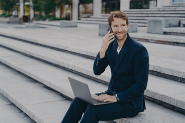 Young attractive businessman working on laptop distantly, speaking on smartphone outside