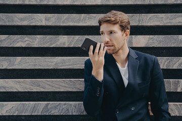 Male executive worker dressed formally holds smartphone near mouth talks on speakerphone