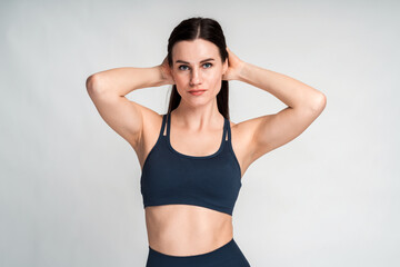 Fototapeta na wymiar Young fit pretty woman with long brunette hair dressed in black sport clothes putting her arms into her head and posing against white blank wall. Bodybuilding and sport concept