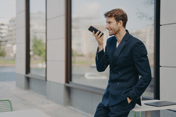 Confident businessman with smartphone using voice assistant app while leaning on cafe table outdoors