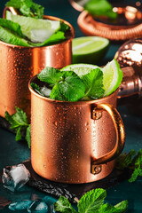 Moscow mule cocktail in copper mug with lime, ice, ginger beer, vodka and mint. Deep blue background and bar tools