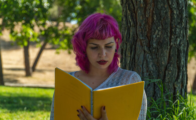 mujer de cabello rosa leyendo un libro junto a un árbol , mujer con un libro amarillo ,  mujer con labios rojos sosteniendo un cuaderno amarillo en la mano - obrazy, fototapety, plakaty
