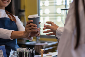 A beautiful Asian barista handing a cup of coffee to customers who wait for orders at the coffee shop counter before chatting and greeting customers before leaving the shop with impressive service.