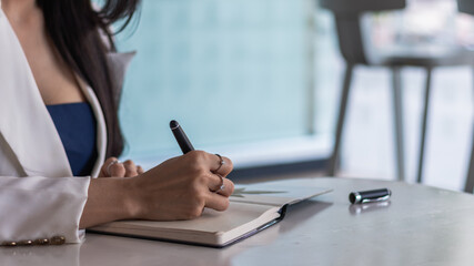 An Asian woman sits at her office desk and uses a pen to write down appointments in her notebook as a reminder. Notebooks are still popular for note taking and are used a lot in school.