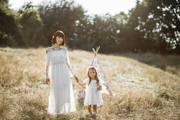 Horizontal outdoor shot of happy stylish family, mom and daughter, dressed in indian boho style with feather in hair, posing in wild meadow at summer sunset. Teepee wigwam behind © sofiko14