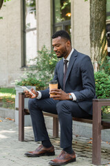 Obraz premium african american businessman using mobile phone and holding coffee to go and sitting on bench