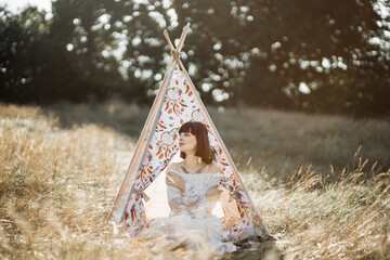 Stylish young hippie woman wearing boho dress and feather hair accessories, sitting near the wigwam tent outside, in beautiful wild summer field © sofiko14