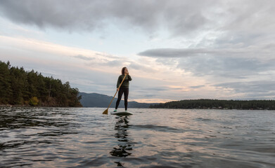 Adventurous White Caucasian Adult Woman Paddle Boarding in the Pacific Ocean during cloudy sunrise. Located near Victoria, Vancouver Island, British Columbia, Canada.