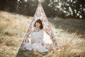 Young woman sitting near teepee tent outdoors in the field. Summer shot of gorgeous likable indian lady in white dress and feathers in hair, sitting on the grass of wild meadow near wigwam © sofiko14