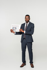 full length of african american man in eyeglasses showing business newspaper isolated on grey