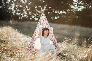 Native American stunning relaxed woman, sitting near stylish boho tee pee wigwam tent in summer field at sunset, looking at the sky and holding her arms behind head © Sofiia Shunkina
