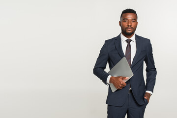 african american businessman in suit holding laptop while standing with hand in pocket isolated on...