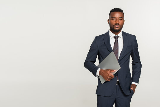 african american businessman in suit holding laptop while standing with hand in pocket isolated on grey