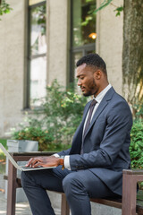 bearded african american businessman typing on laptop while sitting on bench