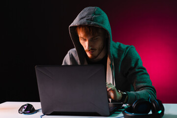 Hacker with a laptop sits at the table, dark background and pink neon light behind, head covered with a hood.