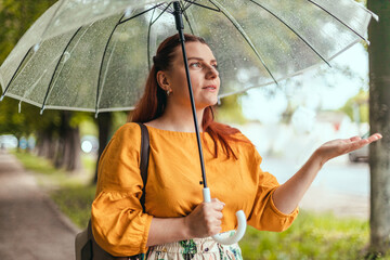 Happy beautiful woman standing under transparent umbrella and stretching hand catching water drops...