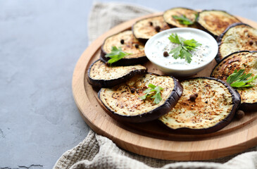 Appetizer of fried eggplant slices with aromatic spices and cream sauce on a wooden plate on a gray background. Side view