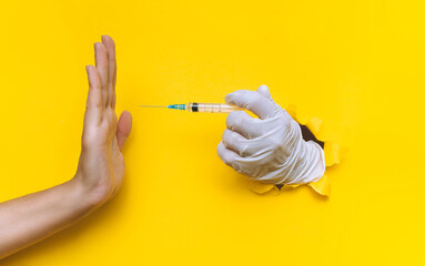 The nurse hand in a white latex glove holds a syringe for vaccination, but collides with a hand in the form of a Stop gesture. Coronavirus anti-vaccination concept.Yellow paper, torn hole, copy space