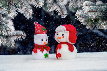 Two toy snowmen under a spruce tree on a dark background in winter during a snowfall. Greeting Christmas and New Year card