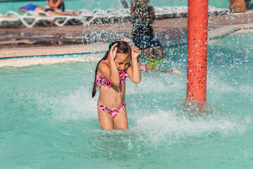 Summer holidays on a sunny day in an outdoor water park. A little girl under the water jets on a...