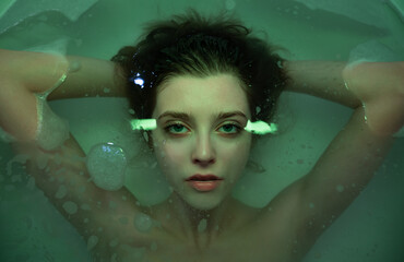 Portrait of young beautiful woman in the bath in turquoise water and colored lights, top view. Copy...