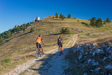 Obraz premium Man and woman riding mtb to castle ruins on mountain top at Rocca Calascio, italian travel destination, Gran Sasso National Park, Abruzzo, Italy. Clear blue sky