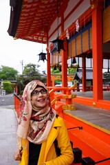 Fototapeta na wymiar Portrait of beautiful young Asian muslim woman wearing hijab and eyeglasses inside Fushimi Inari Shrine in Kyoto. Face smiling and looking up. Happy expression. Outer pavilion in background.