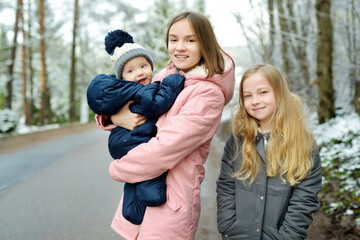 Fototapeta na wymiar Two big sisters and their baby brother having fun outdoors. Two young girls holding their baby boy sibling on winter day. Big age difference between siblings.