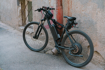 Fototapeta na wymiar Black electric bicycle with battery on the frame stands on the city's street near the wall. Outdoor. Transportation. Electricity. Electric. Ecological. Electric drive. Engine