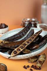 Organic carob pods, seeds in a ceramic plate on a beige background, locust bean healthy food,...