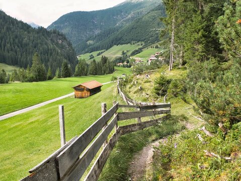 Panoramablick von einem Wanderweg oberhalb der normalen Route &uuml;ber einen Zaun Richtung Gramais in den Alpen in &Ouml;sterreich mit Wanderweg unten, hohen Bergen, H&uuml;tte und Wald