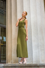 Dressing in a green Maxi Tank Dress, creamy high heels,  crossing legs and one hand touching her shoulder, a young fashion black girl is standing  by a huge column outside an office building, relaxing