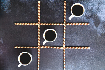 Businessman working morning with cups of hot coffee and sweet croissants on black background. Game of noughts and crosses. Top view, copy space. Flat lay. Creative concept of modern breakfast food. 