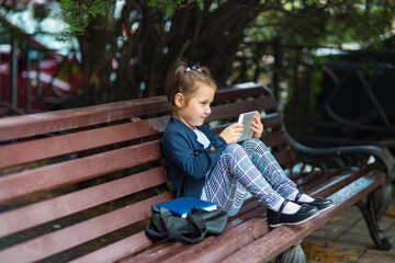 a little girl in a school uniform, sitting on a bench, and holding a tablet in her hands