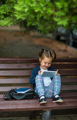 a little girl in a school uniform, sitting on a bench, and holding a tablet in her hands