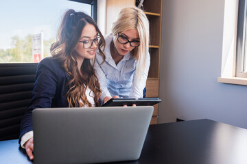 Two young female colleagues looking at tablet screen, satisfied with project results, making new...