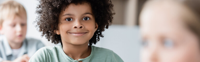 happy african american boy smiling at camera near blurred classmates, banner