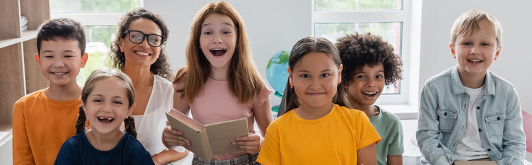 excited girl holding book near happy multiethnic classmates and african american teacher, banner